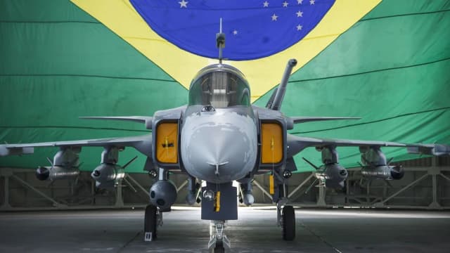 Caça Gripen da Força Aérea Brasileira visto de frente, com a bandeira do Brasil ao fundo