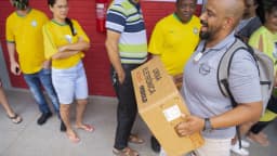 Eleitor carrega caixa de urna eletrônica em fila de seção eleitoral, com outros eleitores ao fundo