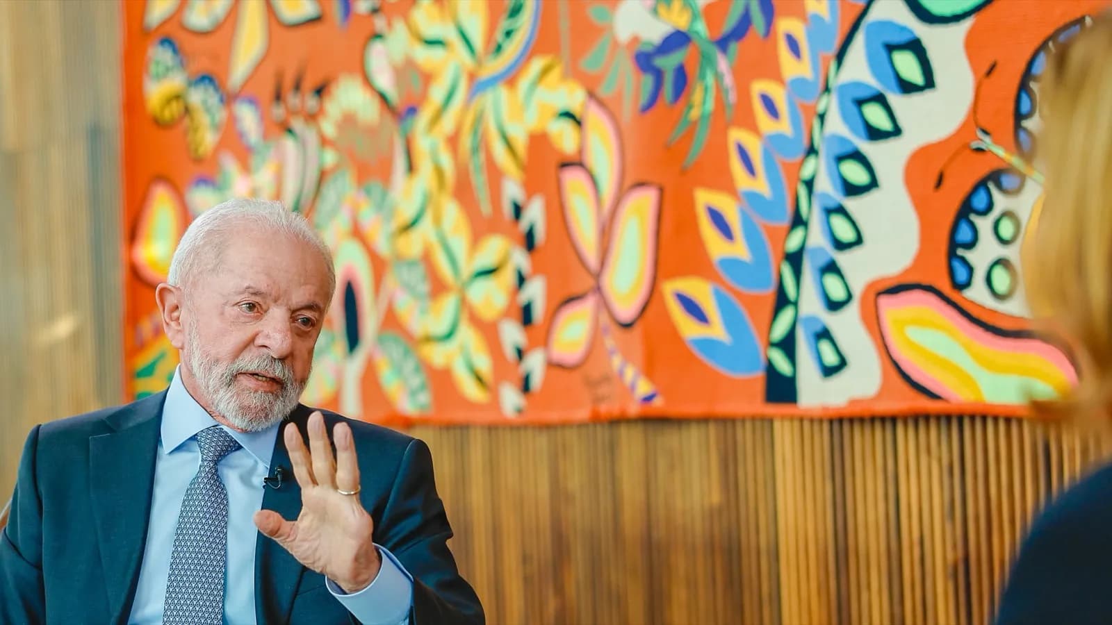 Lula gesticula durante entrevista de televisão, com painel colorido ao fundo