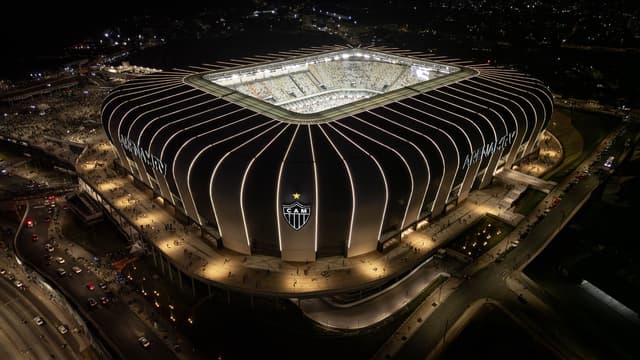 Imagem aérea de arquivo da Arena MRV, estádio do Atlético-MG em Belo Horizonte, iluminada à noite, com o escudo do clube na fachada e torcedores na esplanada