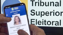 Mão segura celular com o aplicativo e-Título aberto diante de placa do Tribunal Superior Eleitoral