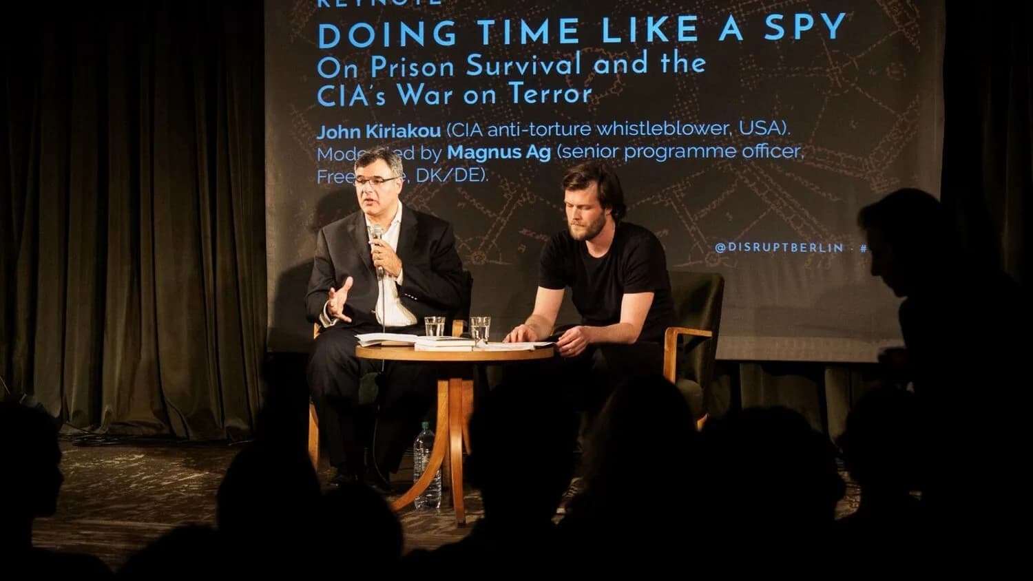 John Kiriakou fala sentado ao lado de um moderador em evento sobre espionagem e tortura da CIA