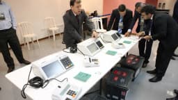 Autoridades inspecionam fileira de urnas eletrônicas dispostas sobre mesa em cerimônia de auditoria