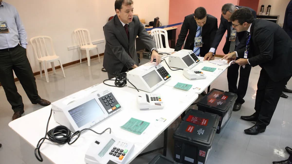 Autoridades inspecionam fileira de urnas eletrônicas dispostas sobre mesa em cerimônia de auditoria