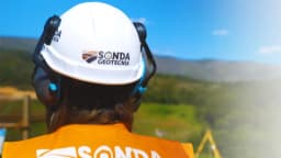 Profissional com capacete e colete da Sonda Geotecnia observa área de trabalho em campo, com serra ao fundo