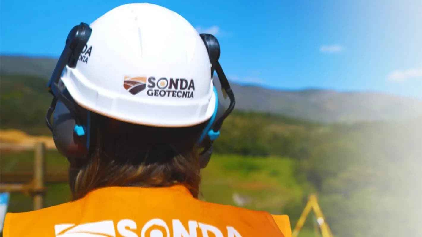 Profissional com capacete e colete da Sonda Geotecnia observa área de trabalho em campo, com serra ao fundo