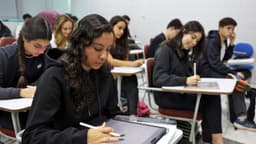 Estudantes sentados em carteiras de uma sala de aula, escrevendo em cadernos e tablets, em imagem de arquivo