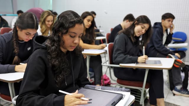 Estudantes sentados em carteiras de uma sala de aula, escrevendo em cadernos e tablets, em imagem de arquivo