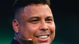 Ronaldo Nazário sorri durante evento, usando microfone de lapela e terno escuro