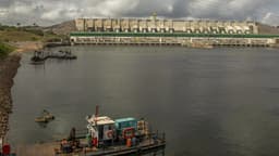 Barragem de usina hidrelétrica com reservatório em primeiro plano e embarcação de apoio, sob céu nublado; imagem de arquivo