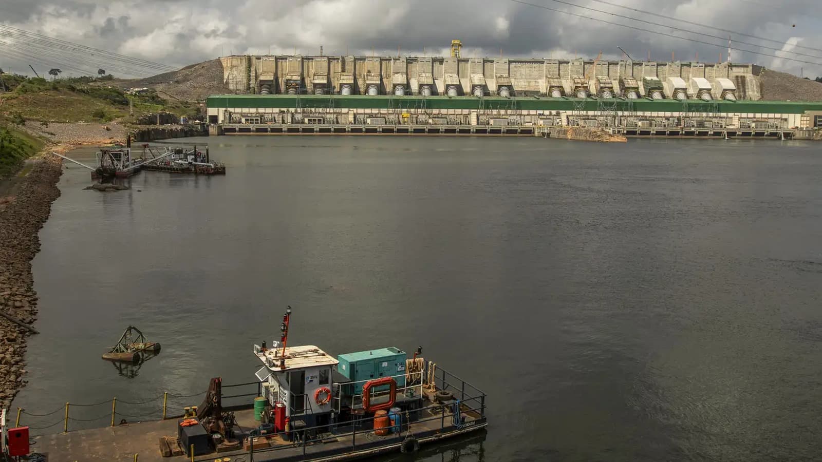 Barragem de usina hidrelétrica com reservatório em primeiro plano e embarcação de apoio, sob céu nublado; imagem de arquivo