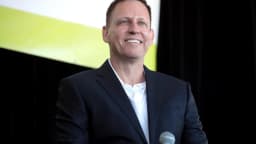 Peter Thiel sorri durante evento, de terno escuro, ao lado de um microfone