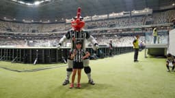 Mascote do Atlético-MG, o Galo Doido, ao lado de uma criança no gramado da Arena MRV lotada