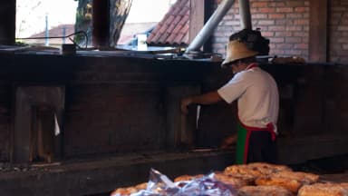 Padeiro trabalha em forno tradicional de pão, com pães recém-assados em primeiro plano
