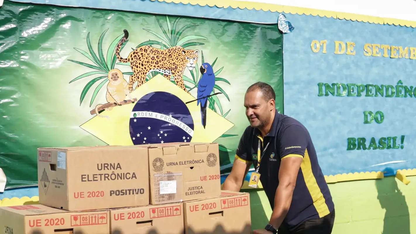 Agente eleitoral com urnas eletrônicas em frente a painel com bandeira do Brasil