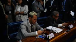 Cleitinho Azevedo sentado em mesa de comissão no Senado, olhando para o celular