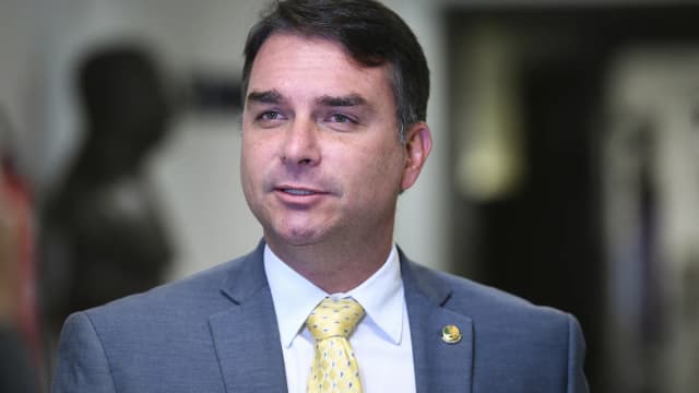 Flávio Bolsonaro fala a entrevistadores nos corredores do Senado, vestindo terno cinza e gravata amarela