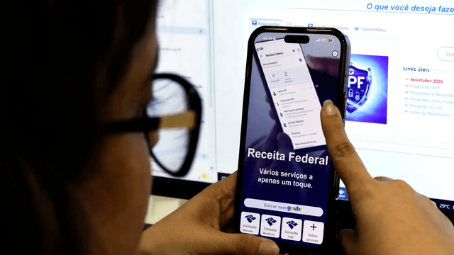 Foto de arquivo: pessoa vista de costas segura celular com o aplicativo da Receita Federal aberto, diante de um computador com a página do Imposto de Renda