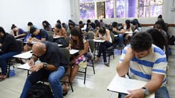 Estudantes adultos sentados em carteiras de uma sala de aula durante a aplicação de uma prova; imagem de arquivo