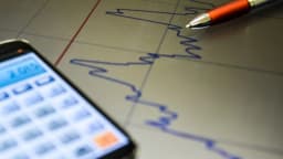 Gráfico de mercado financeiro em tela de celular ao lado de caneta sobre relatório impresso