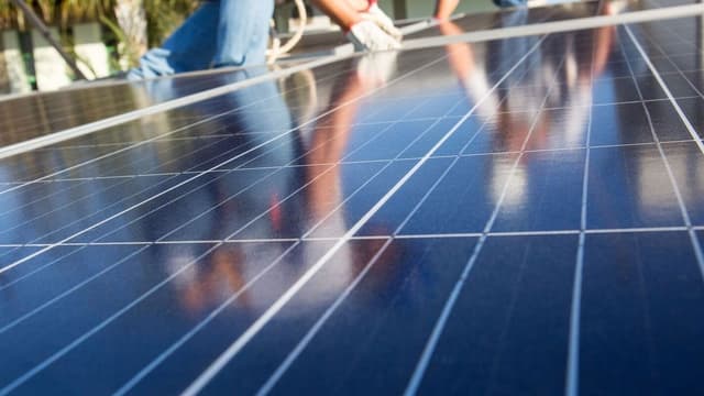 Técnico com luvas instala painéis solares fotovoltaicos, vistos de perto e refletindo o céu