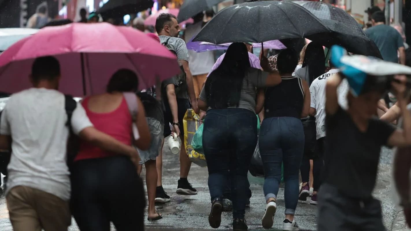 Pessoas caminham sob chuva forte protegidas por guarda-chuvas em via urbana (foto de arquivo, ilustrativa)