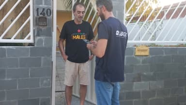 Recenseador do IBGE, identificado por camisa do instituto, entrevista morador em porta de casa
