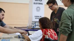 Eleitora idosa é auxiliada por mesários em cabine de votação