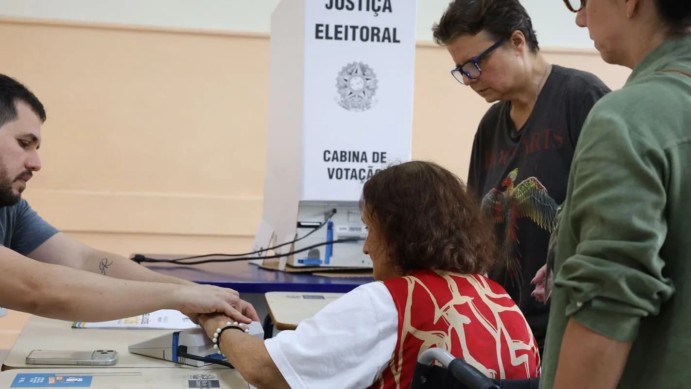 Eleitora idosa é auxiliada por mesários em cabine de votação
