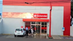 Fachada de uma agência do banco Santander, com letreiro vermelho e branco da marca sobre a porta de vidro e clientes na calçada. Foto de arquivo.