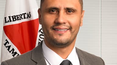Retrato oficial do senador Cleitinho, de terno cinza, sorrindo, com a bandeira de Minas Gerais ao fundo
