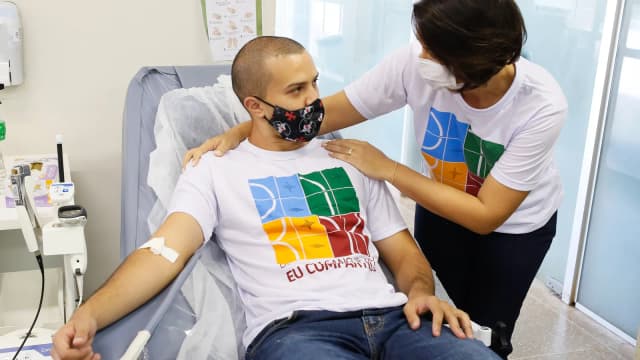 Homem doando sangue em hemocentro, deitado em maca, com profissional de saúde ao lado
