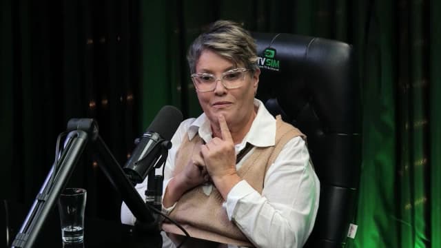 Cássia Barbosa, com a mão no queixo, fala pensativa durante entrevista no estúdio da TV Sim Brasil
