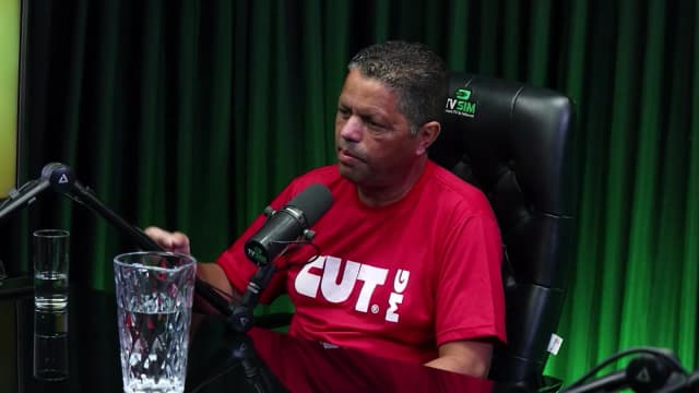 Jairon Nogueira Filho, presidente da CUT em Minas Gerais, de camiseta vermelha da CUT-MG, fala gesticulando em estúdio com cortina verde ao fundo, ao lado de um copo d'água na mesa.