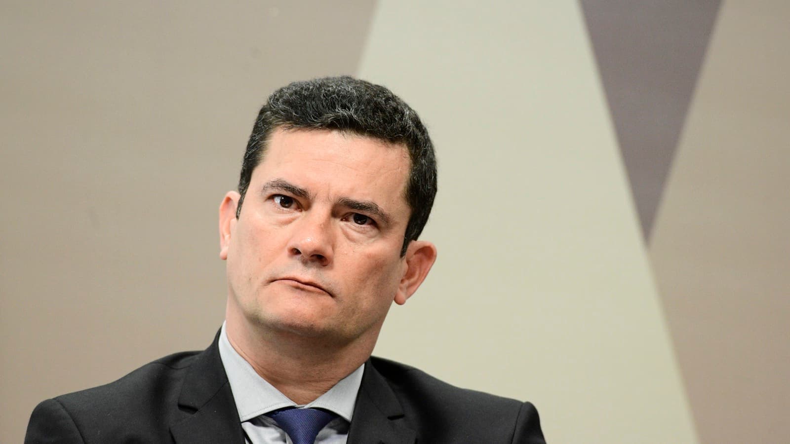 Retrato de Sérgio Moro em plano fechado, de terno escuro e gravata azul, com expressão séria e fundo neutro do Senado. Imagem de arquivo.