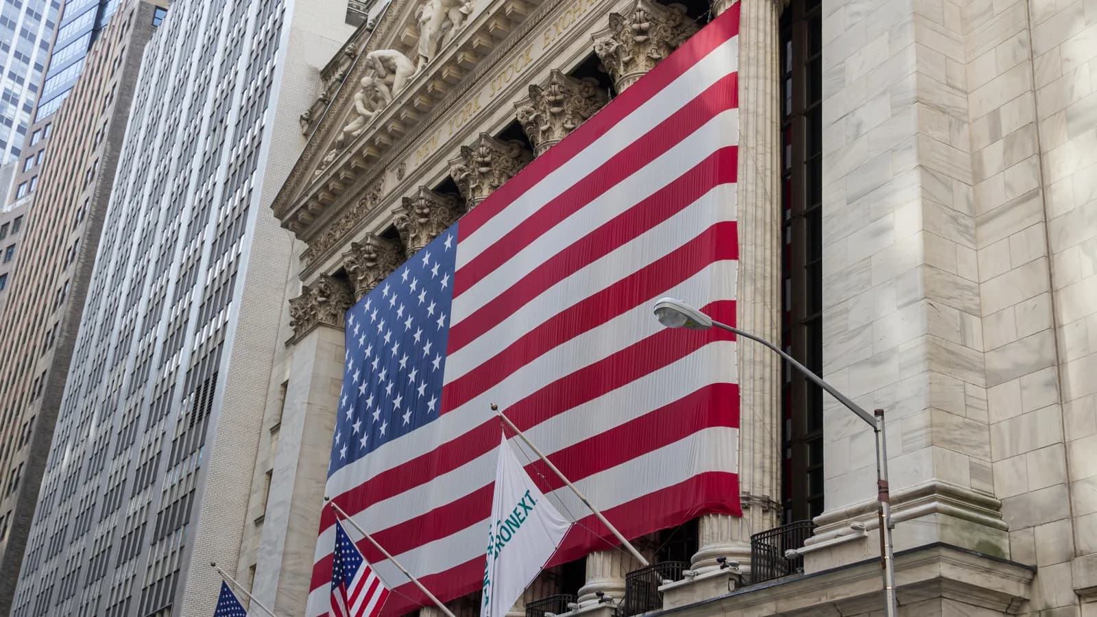 Fachada da Bolsa de Nova York (NYSE) em Wall Street, com uma grande bandeira dos Estados Unidos estendida entre as colunas