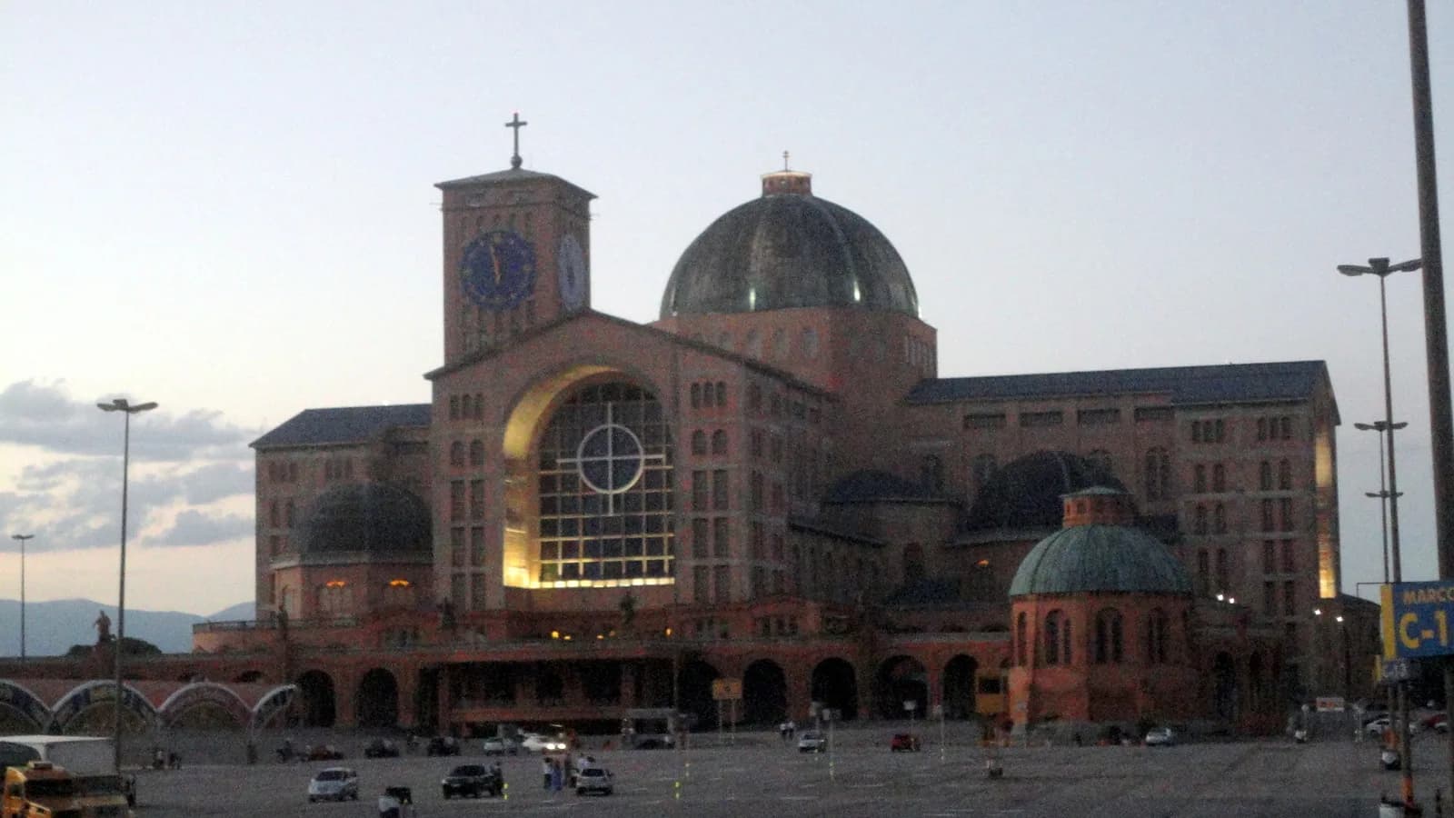 Fachada da Basílica Nacional de Nossa Senhora Aparecida ao entardecer, com esplanada e carros estacionados