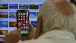 Homem segura um iPhone com a tela de aplicativos aberta, com um computador ao fundo