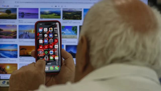 Homem segura um iPhone com a tela de aplicativos aberta, com um computador ao fundo