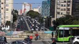 Entregador de aplicativo em moto e ciclista de entrega no trânsito de avenida movimentada em São Paulo, com ônibus e carros ao fundo