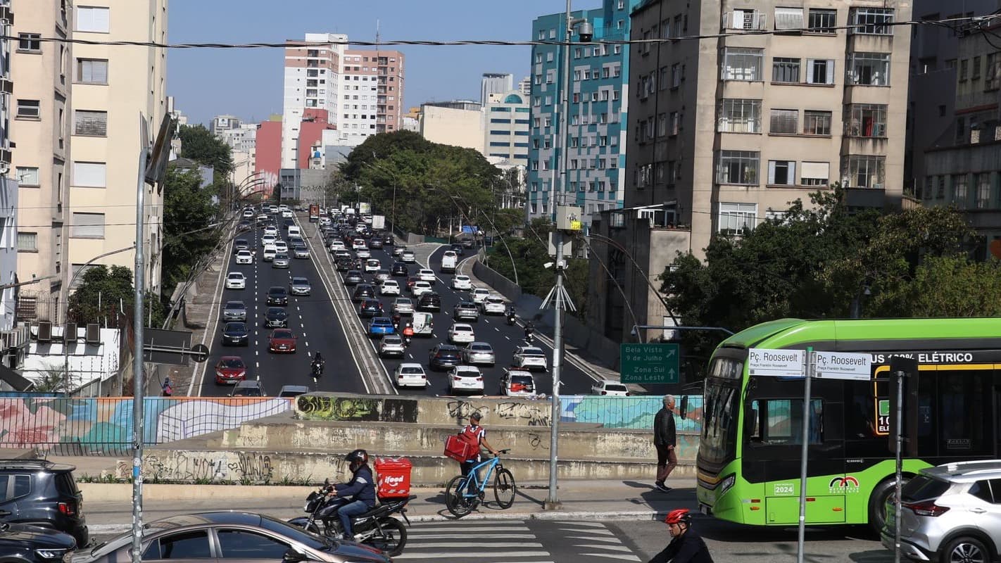 Entregador de aplicativo em moto e ciclista de entrega no trânsito de avenida movimentada em São Paulo, com ônibus e carros ao fundo