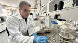 Pesquisador de jaleco branco e luvas manuseia amostra em laboratório com bancada de granito e vidrarias