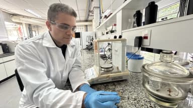 Pesquisador de jaleco branco e luvas manuseia amostra em laboratório com bancada de granito e vidrarias