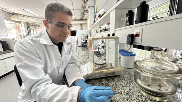 Pesquisador de jaleco branco e luvas manuseia amostra em laboratório com bancada de granito e vidrarias
