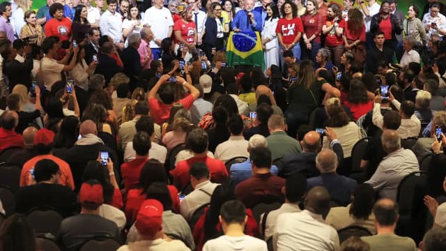 O presidente Luiz Inácio Lula da Silva discursa ao microfone, segurando uma bandeira do Brasil, diante de apoiadores