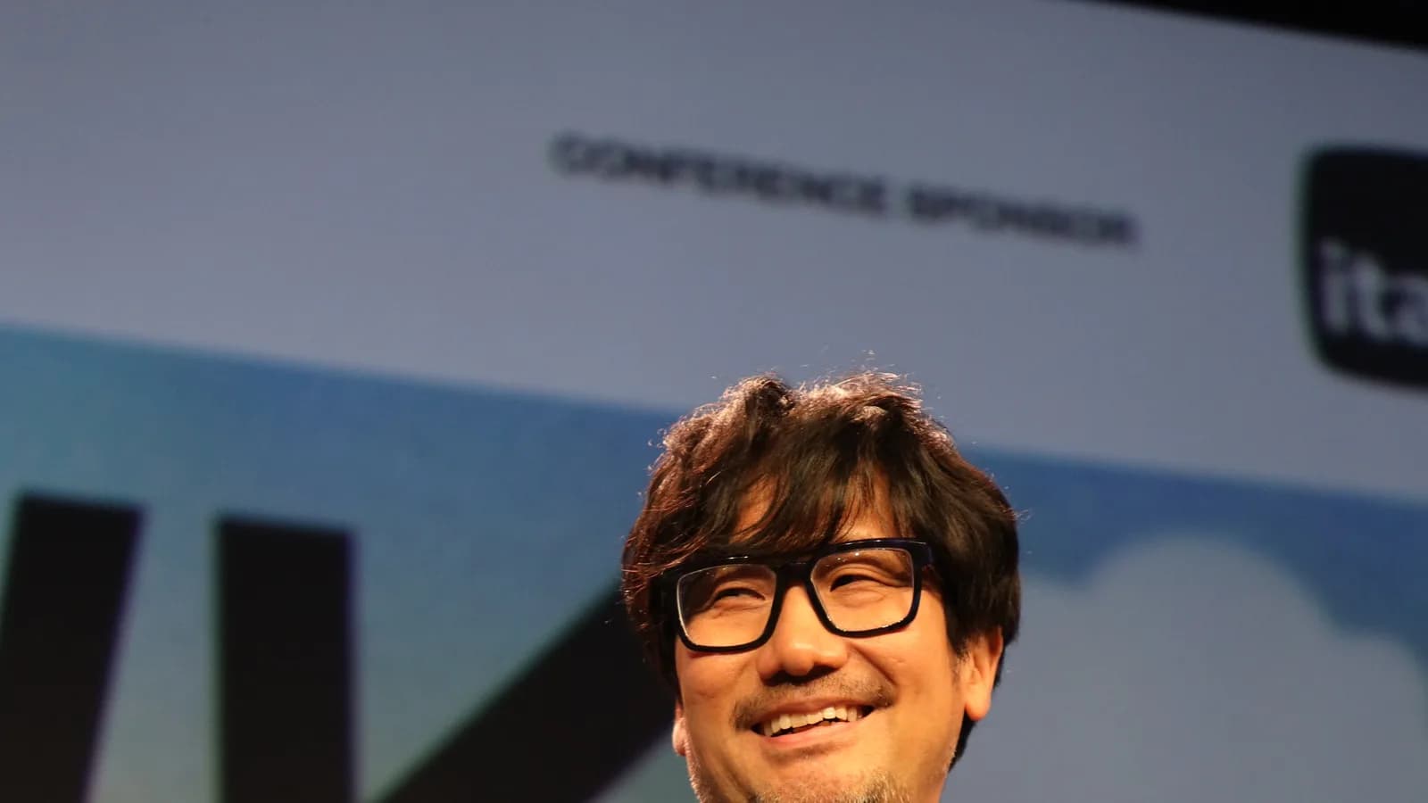 Hideo Kojima sorrindo em evento público, usando óculos e camiseta estampada com arte de Death Stranding