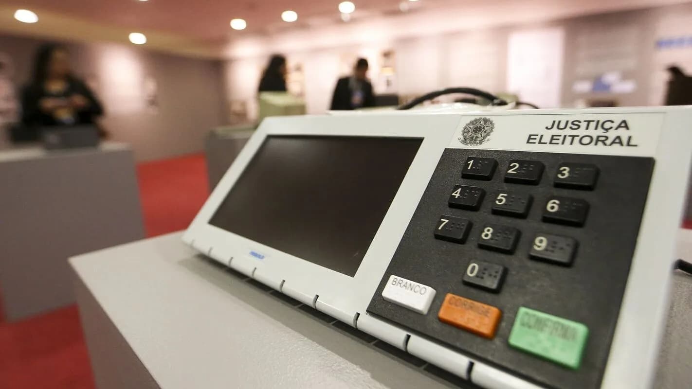 Urna eletrônica da Justiça Eleitoral em primeiro plano, com pessoas ao fundo em sala de votação