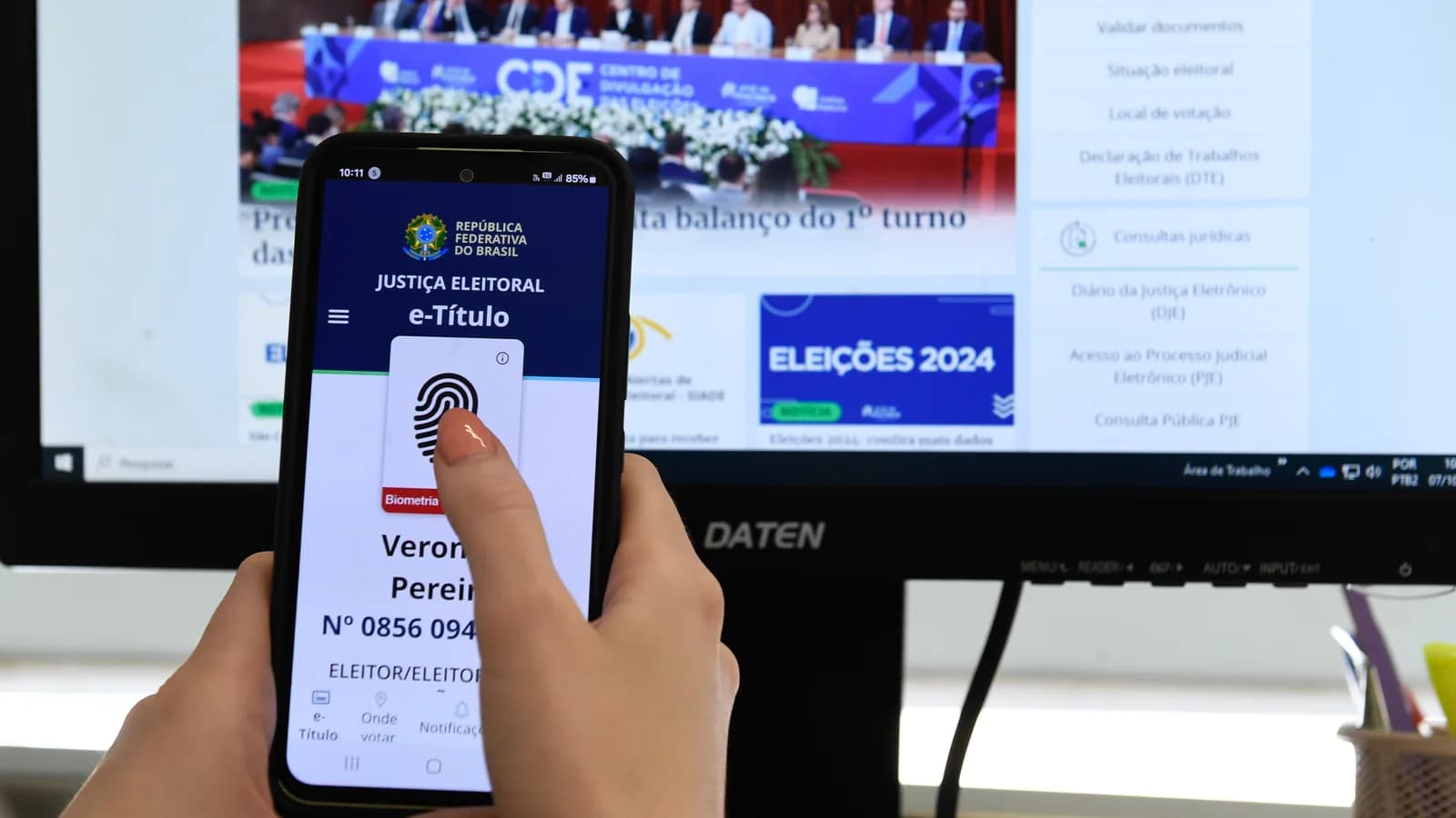 Mão segura celular com o aplicativo e-Título aberto, com evento oficial da Justiça Eleitoral ao fundo
