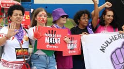Imagem de arquivo do ato do 8 de março de 2026: mulheres em manifestação de rua seguram cartazes vermelhos com a frase Parem de nos matar e uma faixa com a palavra Mulheres