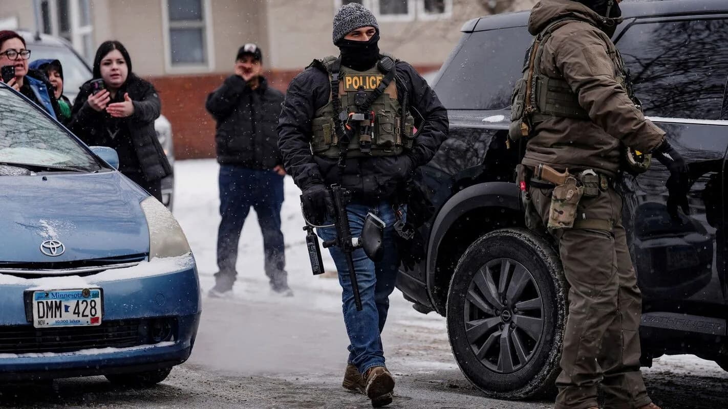 Agentes do ICE em capacete e colete tático perto de um veículo, com pessoas observando ao redor